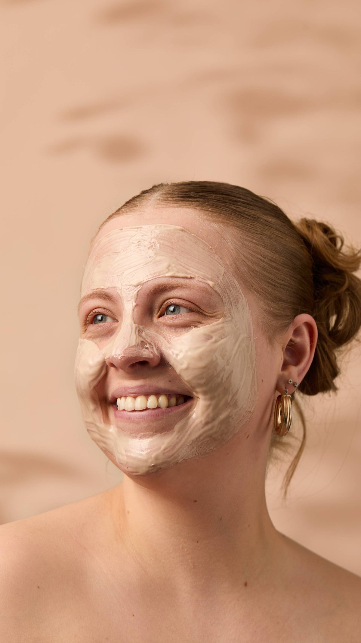 An image of the model's face on a warm, beige background. Their copper hair is tied back, and they have applied the thick Coconut Shie face mask over their full face as they smile.