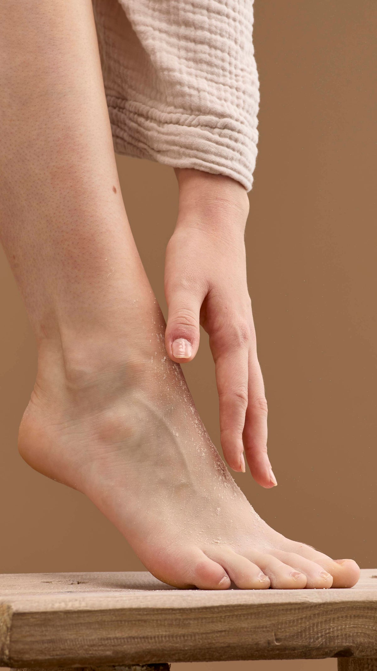 The image shows a hand rubbing a pale cream 'T For Toes' foot powder onto a bare foot and ankle. The foot rests on a wooden stool, with the person's robe visible against a solid beige background.