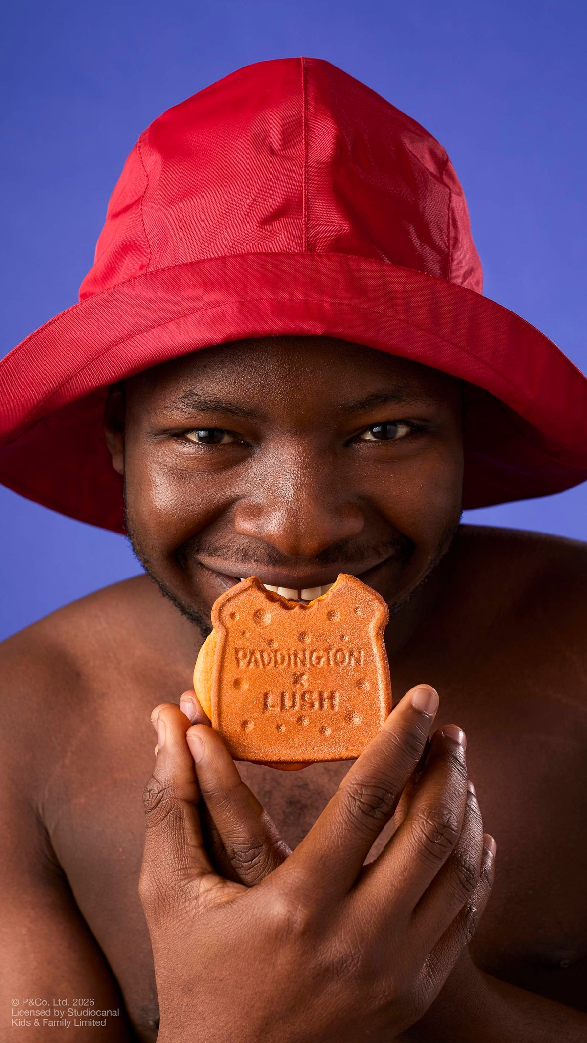 A model in a red rain hat smiles directly at the camera while holding a "Marmalade Sandwich" bubbleroon bubble bar to their mouth, as if about to take a bite. The bubble bar features the text 'PADDINGTON x LUSH' against a solid purple background.