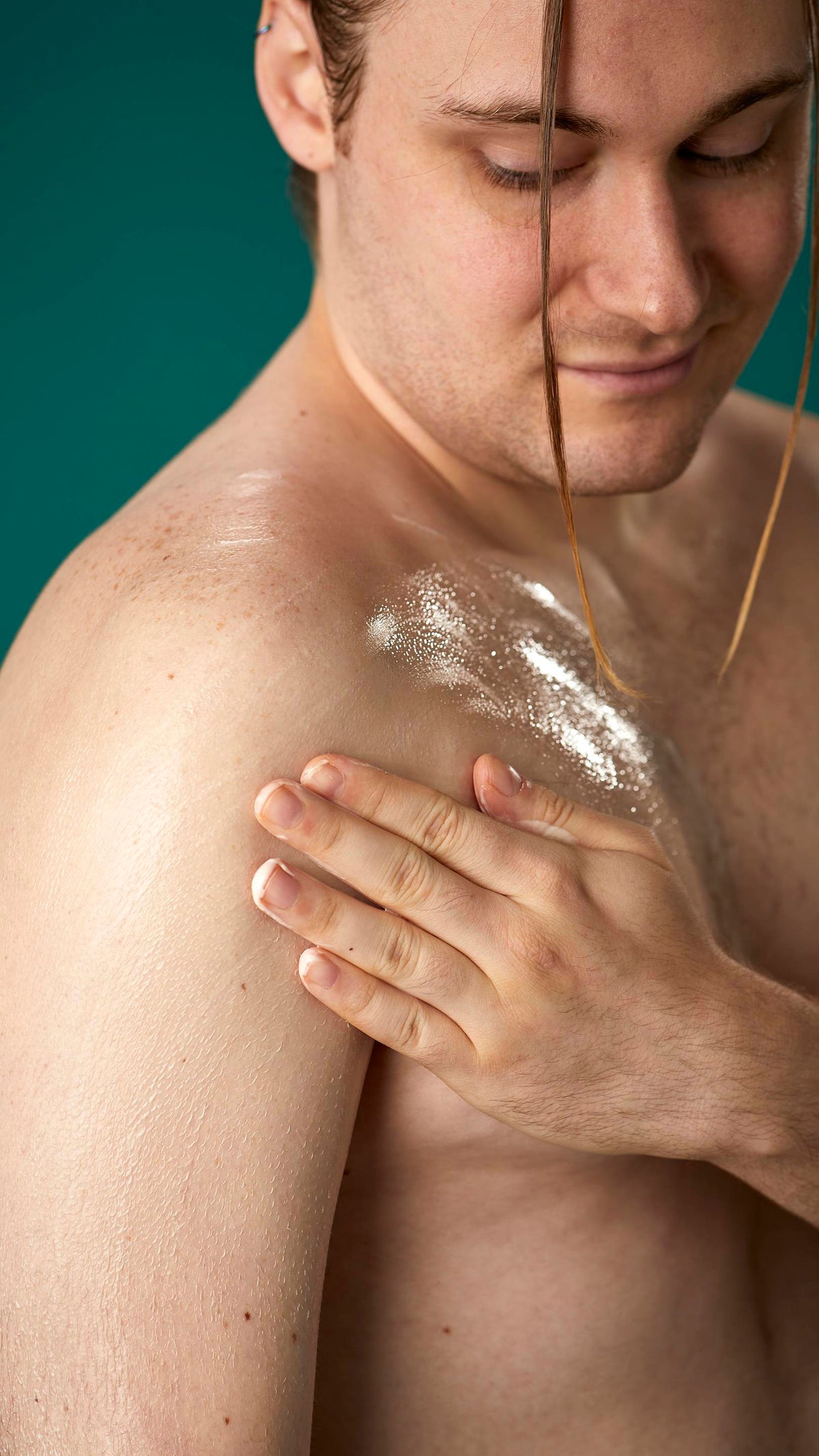 The image shows a topless model on a teal background. They are gently soothing the Braziliant body butter in circular motions over their chest and upper arm.