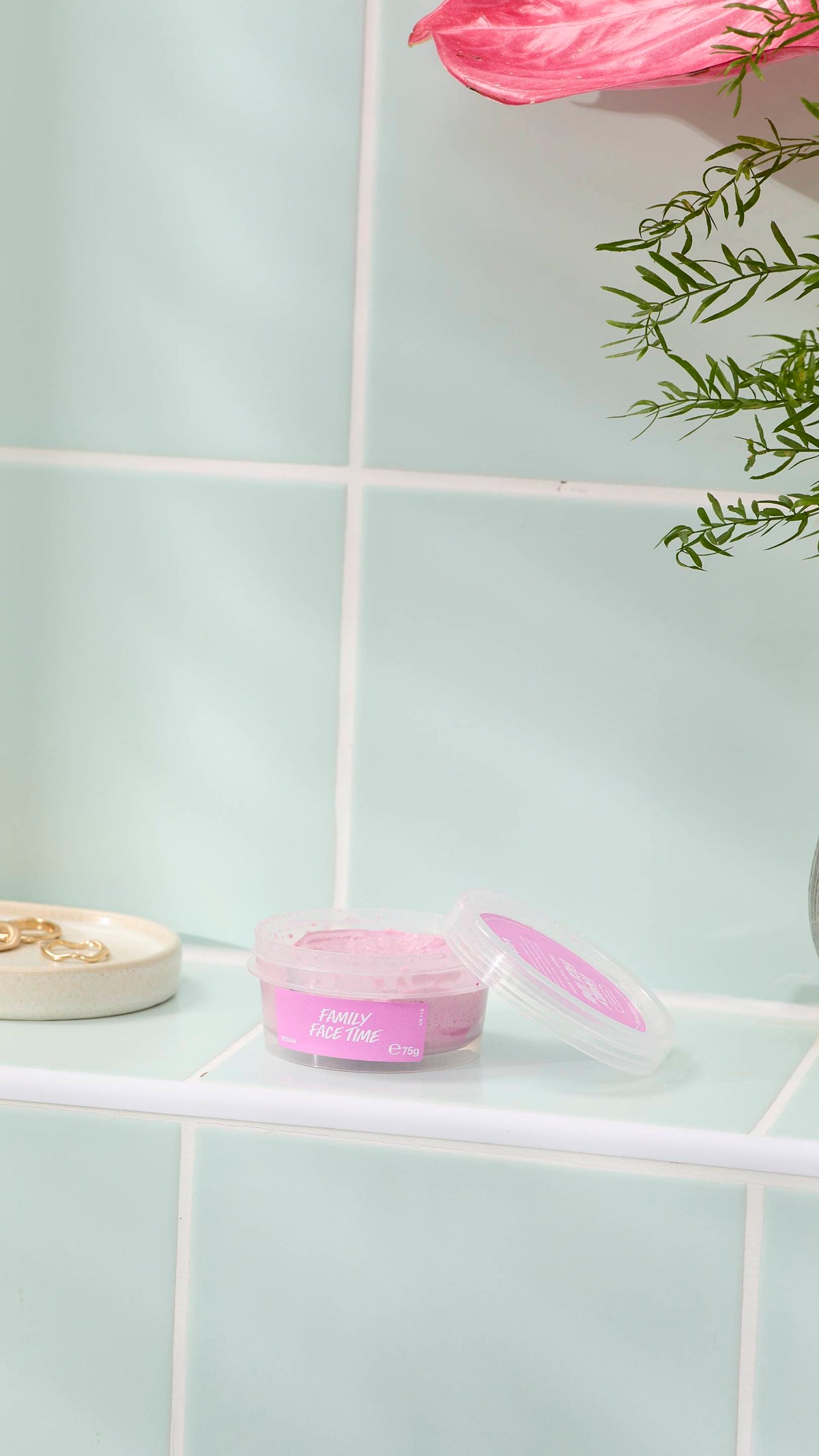 An open, clear plastic pot of 'FAMILY FACE TIME' fresh face mask sits on a pale green tiled ledge. The pot contains a thick, grainy pink face mask, with some residue on the lid. A small dish with gold rings and a pink flower are nearby.