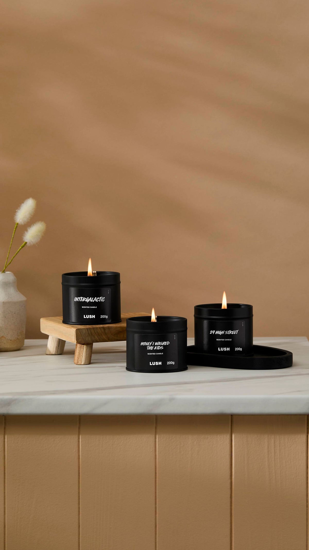 The image shows a warm beige background with a ceramic vase of Hare's Tail Grass. Three back tin candles are sitting with their wicks lit on a marble-topped counter.
