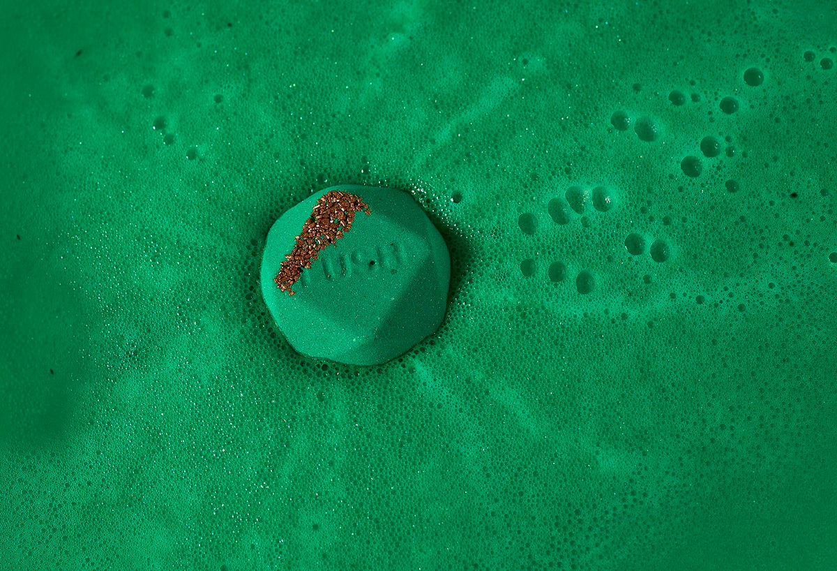 Kokage bath bomb floats in deep, emerald green sea of foam with orange-coloured salt and "Lush" embossed on top.
