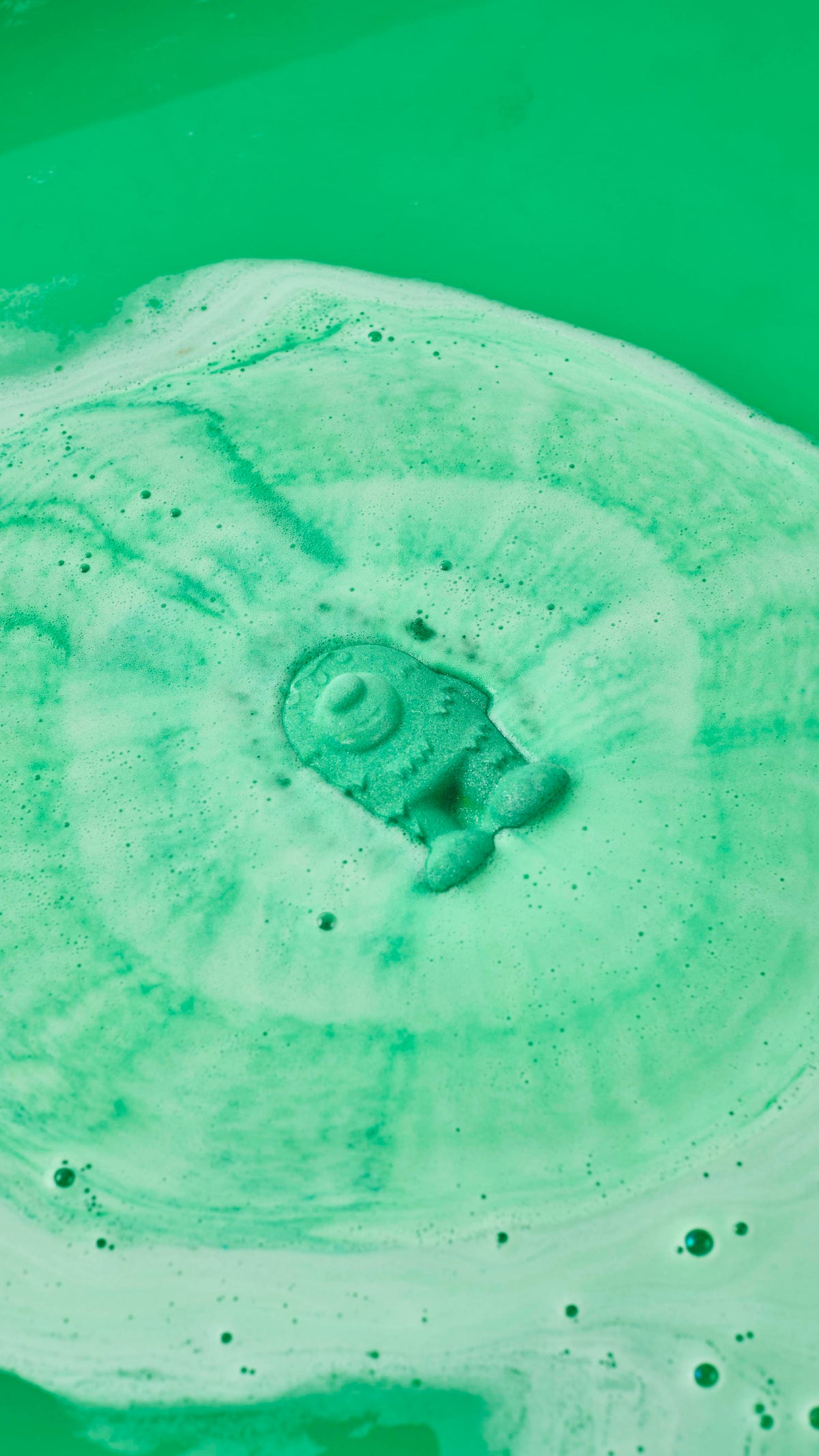 A mint-green, monster-shaped bath bomb dissolves at the centre of a bath, generating concentric ripples of lime-green and pale-green foam. The surrounding water is a vivid, opaque shade of green.